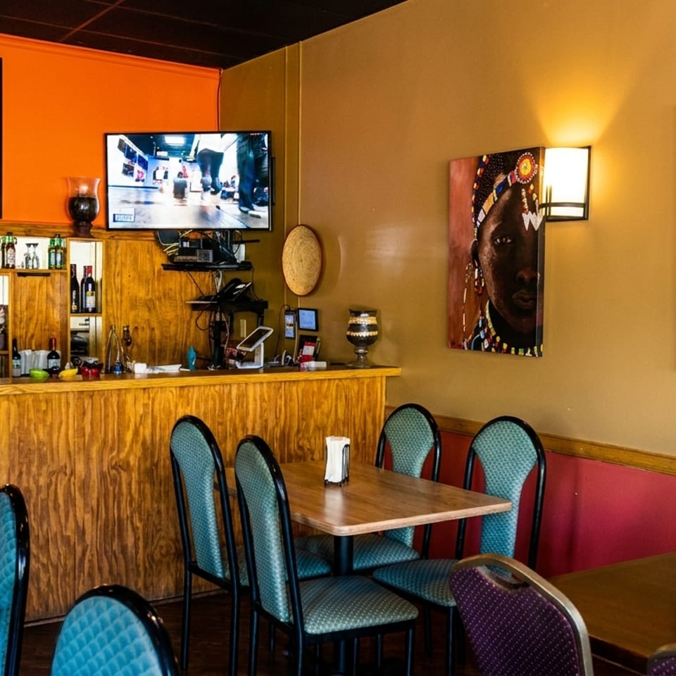 Restaurant interior with wooden bar, tables with teal chairs, and vibrant African-themed artwork on the walls.