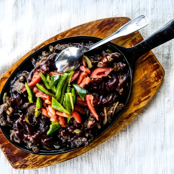 Saucy meat dish with green peppers and tomatoes in a black skillet on a wooden board.