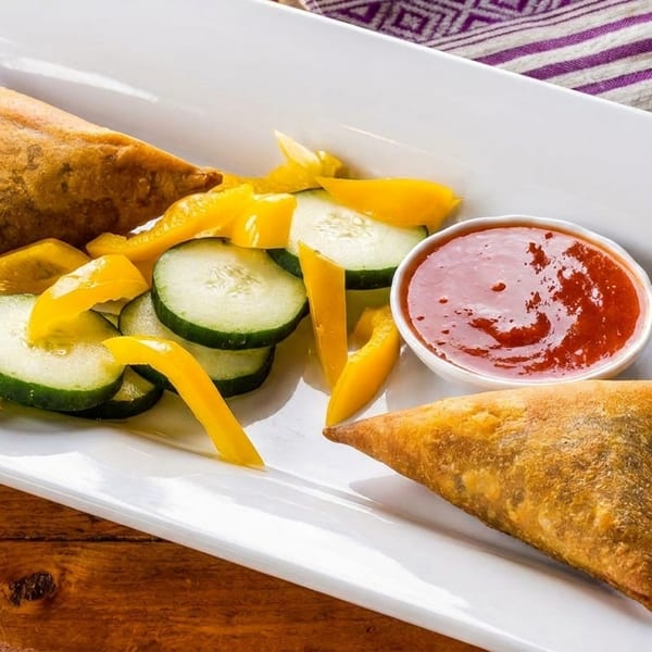 Two golden samosas with sliced cucumbers, yellow bell peppers, and a side of red sauce.